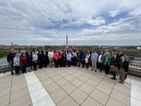 A large group of people stands together on a rooftop terrace with the Washington Monument and surrounding cityscape visible in the background under a partly cloudy sky.