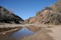 A wash with rocky walls on either side. A standing pool of water at the bottom of the wash reflects part of the rock walls.