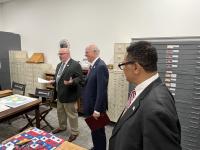 Three people stand together in a room with map drawers and archival furniture. One person speaks while holding papers, and the others listen attentively. Colorful documents and historical materials are laid out on nearby tables.