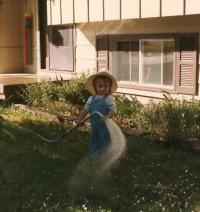 A small child standing in a yard, wearing overalls and a wide-brimmed hat, holding a garden hose that sprays water across the grass near a house.