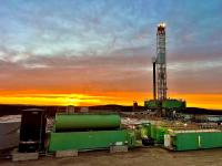An onshore oil and gas drilling rig stands against a vibrant orange and blue sunrise sky. In the foreground, large green industrial mud tanks and a vertical black storage tank sit on a dirt lot. The tall metal derrick tower rises on the right side of the frame with its working lights illuminated.