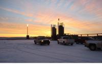 Pickup trucks parked on a snow-covered work site at sunrise, with tall industrial tanks and a gas flare silhouetted against an orange and pink sky.