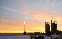 Pickup trucks parked on a snow-covered work site at sunrise, with tall industrial tanks and a gas flare silhouetted against an orange and pink sky.