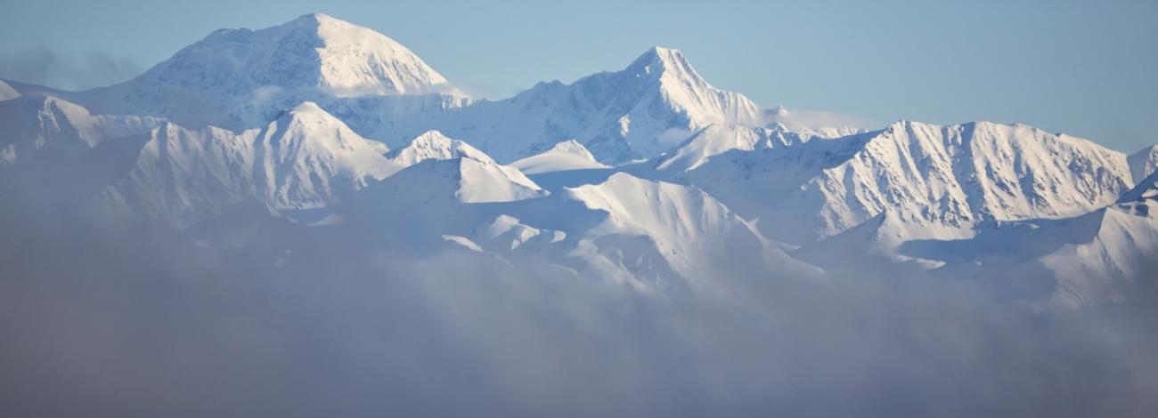 A range of snow-covered mountain peaks rises above a layer of clouds or fog. The rugged, jagged summits are blanketed in snow under a clear pale blue sky. The lower slopes fade into the soft mist below.