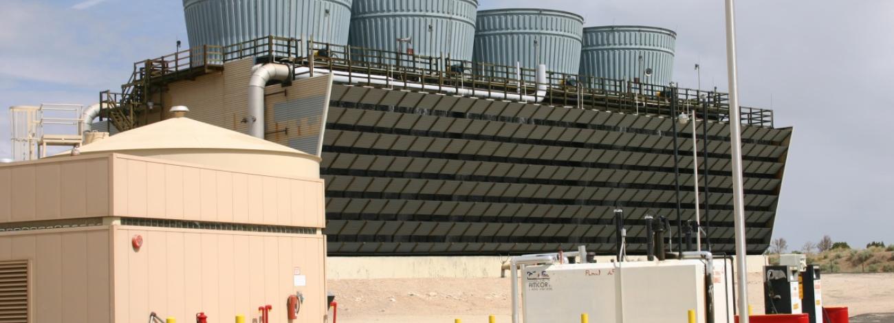 An industrial geothermal facility with large cylindrical cooling towers sits behind a sloped metal structure, with smaller beige utility buildings and equipment in the foreground under a cloudy sky.