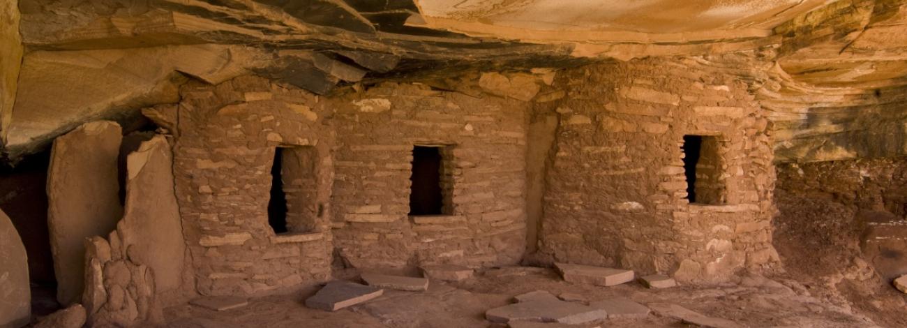 A row of ancient stone dwellings built into a sandstone alcove, with small rectangular doorways and stacked rock walls beneath an overhanging cliff.