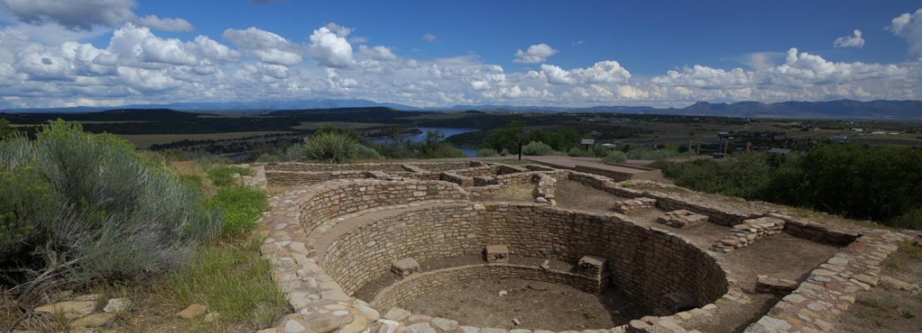 Ancient stone ruins, including a large circular structure (kiva) with masonry walls, sit on a hilltop overlooking a wide valley. A reservoir or lake and distant mountains are visible in the background. The site is surrounded by sagebrush and grasses under a blue sky with scattered white clouds.