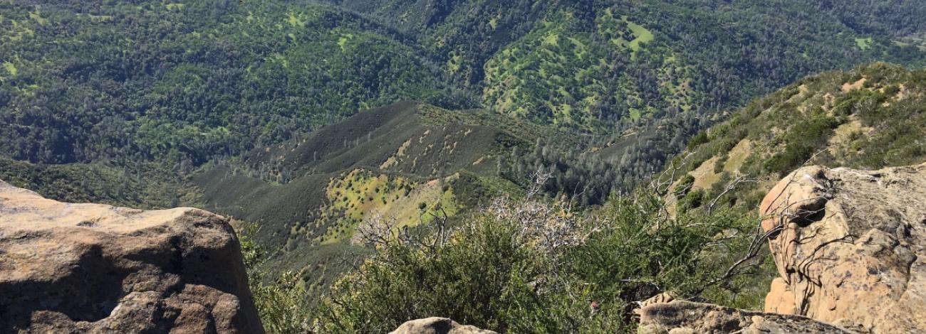 A sweeping view from a rocky overlook shows rolling hills and steep ridges covered in dense green vegetation, with patches of sunlight highlighting different areas of the landscape.
