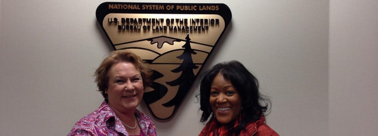 Two people stand in front of a large Bureau of Land Management wall emblem that features a stylized landscape with mountains, trees, and a river. The emblem reads “National System of Public Lands – U.S. Department of the Interior, Bureau of Land Management.”