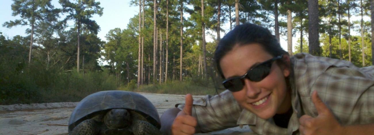 A smiling person wearing sunglasses and a plaid shirt gives two thumbs up while posing next to a tortoise on a sandy path. They are lying low to the ground at the tortoise's level. Behind them is a forest of tall trees and green undergrowth under a bright sky.