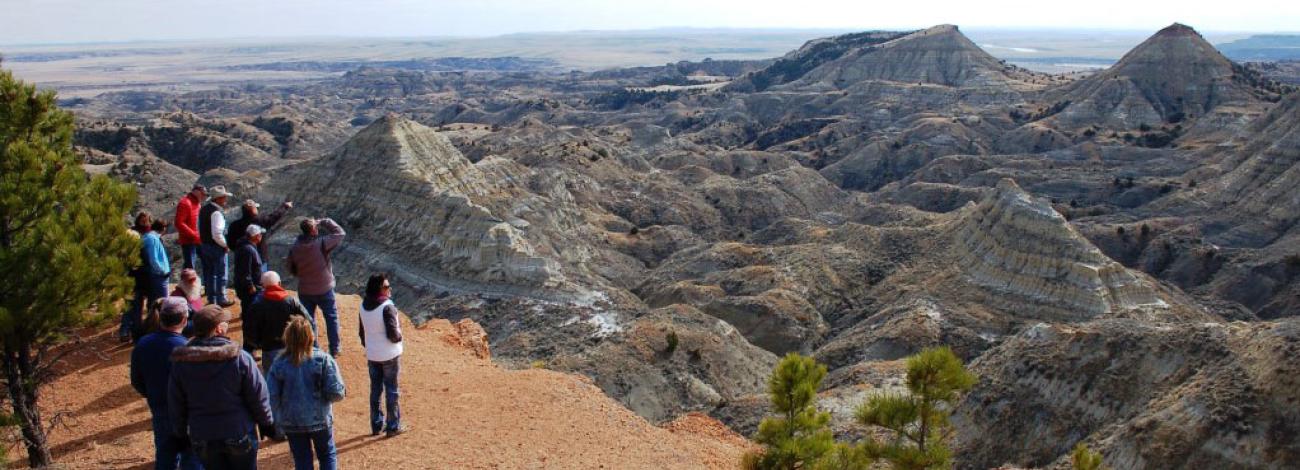 A group of people gathered on a reddish dirt overlook gaze out at an expansive badlands landscape. The terrain features eroded, layered rock formations and ridges in shades of gray and tan, stretching toward flat plains in the distance. A few trees grow near the viewpoint under a hazy sky.