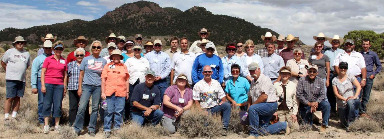A large group of people poses together outdoors in a desert landscape, standing and kneeling among low shrubs with rugged hills and partly cloudy skies in the background.