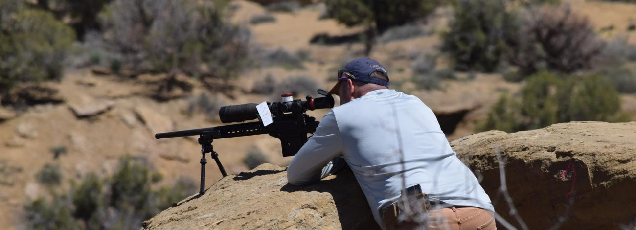 A photo of a man leaning on a rock in the desert, aiming a firearm into the distance.