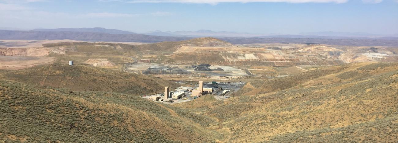 In the foreground we see hills covered with green sagebrush. In the middle distance we see some tan-colored buildings on a gravel pad. In the far distance we see the terraced landscape of an open pit mine under a blue sky with a few white clouds.