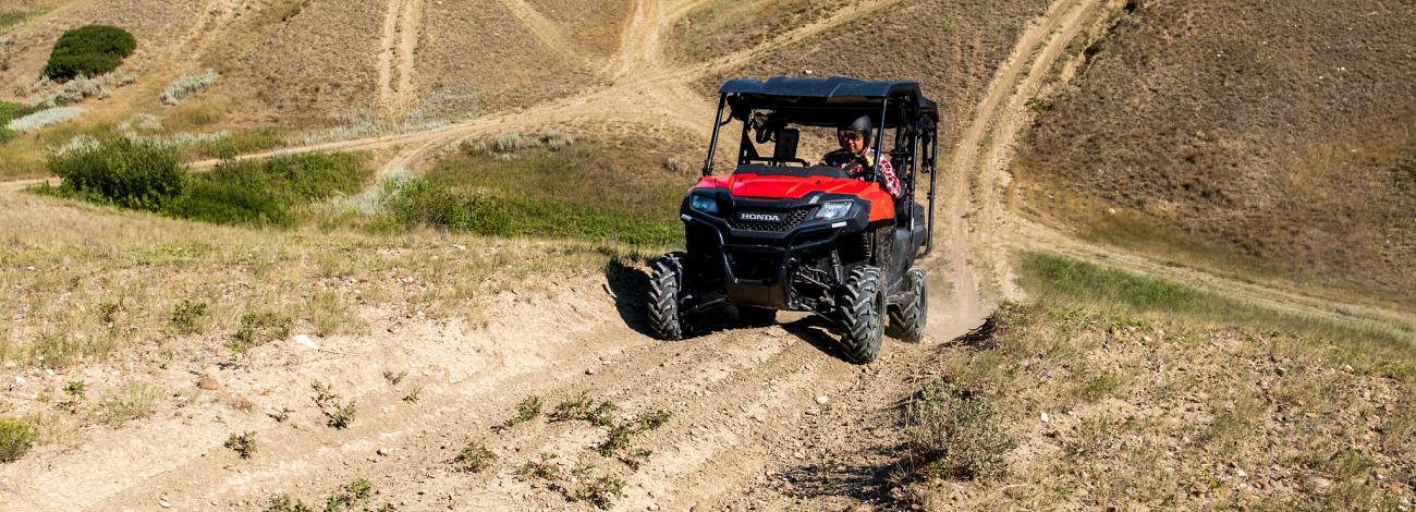 Red off-road utility vehicle driving on a dusty dirt trail through dry, hilly terrain under a clear sky.