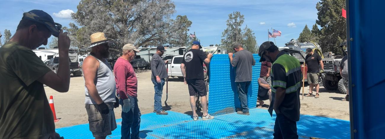 Group of people working outdoors on installing large blue interlocking panels on sandy ground near parked vehicles and trailers, with trees and flags visible in the background under a clear sky.