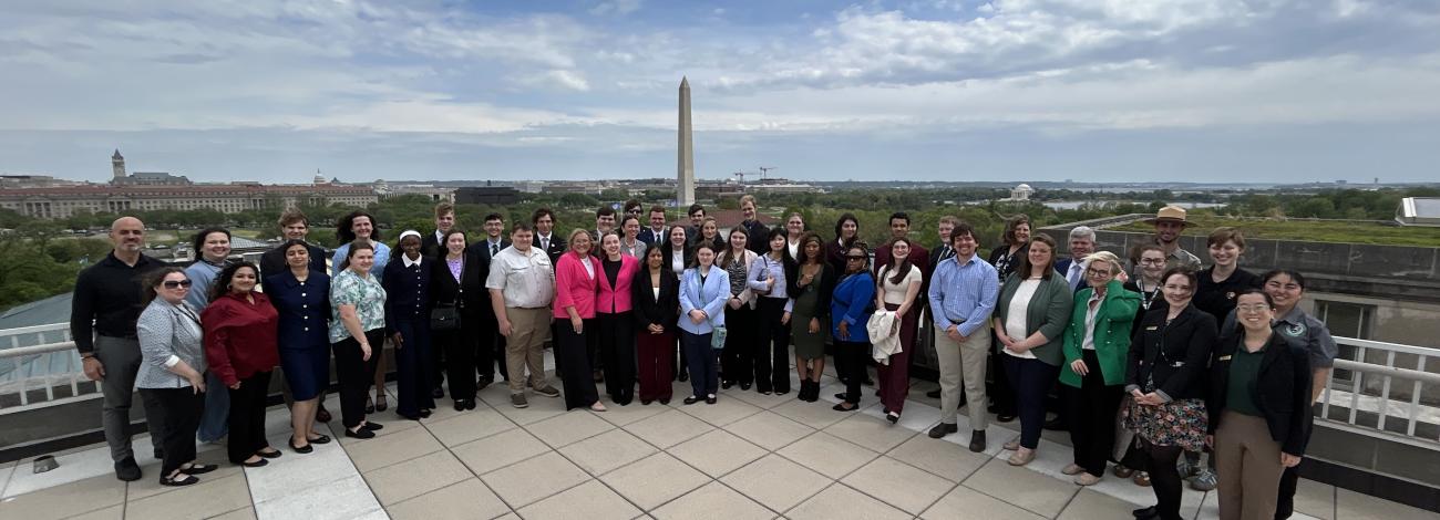 A large group of people stands together on a rooftop terrace with the Washington Monument and surrounding cityscape visible in the background under a partly cloudy sky.