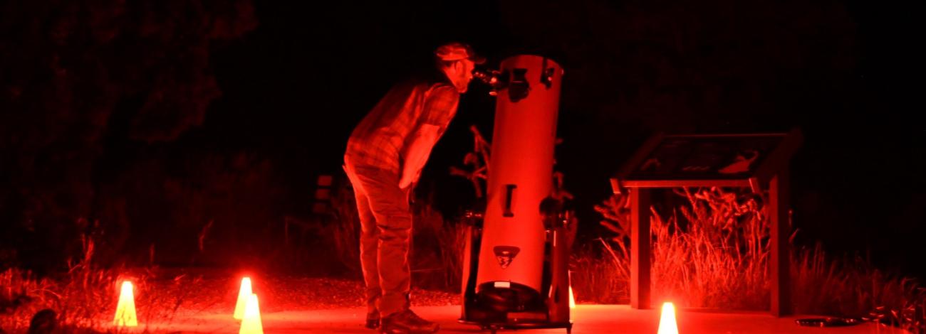 Man gazes through a large telescope, surrounded by red lights.