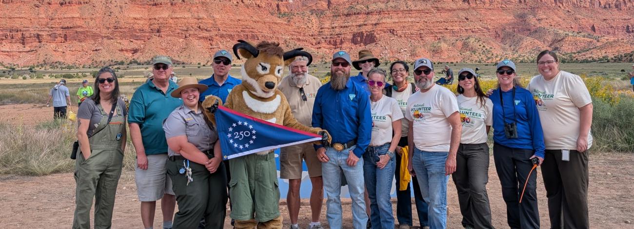 a group of people with an antelope mascot stand in front of distant red-rock cliffs