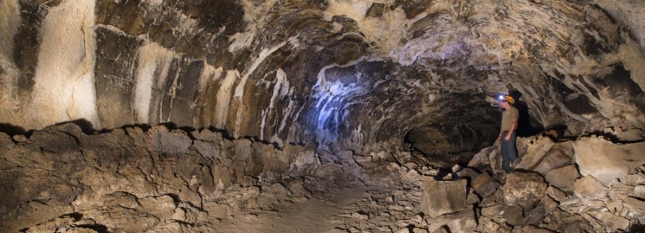 A person wearing a headlamp stands inside a large cave, illuminating the textured rock walls and ceiling. The cave's interior shows streaks of pale mineral deposits against the dark brown rock, with a rocky, uneven floor. The person, standing on the right, gives a sense of the cave's vast scale.