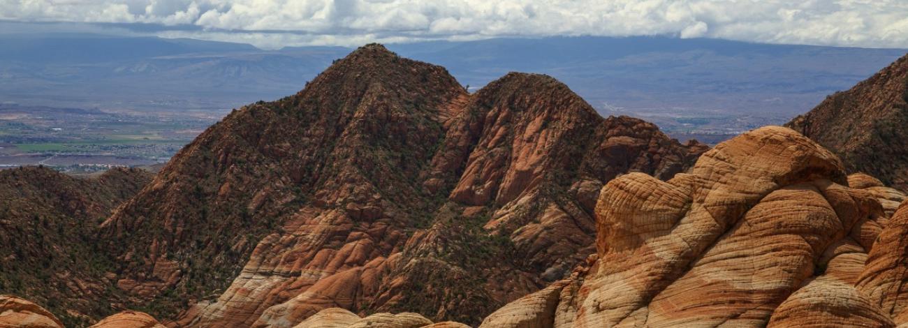 Layered red and beige formations in the foreground with rugged mountain peaks under a cloudy sky in the background.