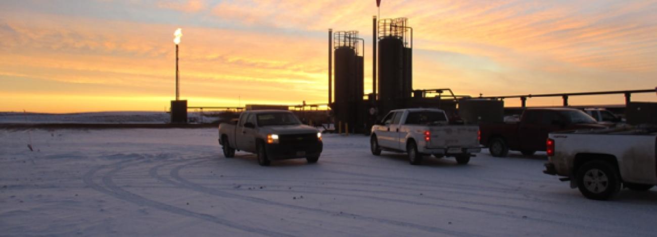Pickup trucks parked on a snow-covered work site at sunrise, with tall industrial tanks and a gas flare silhouetted against an orange and pink sky.