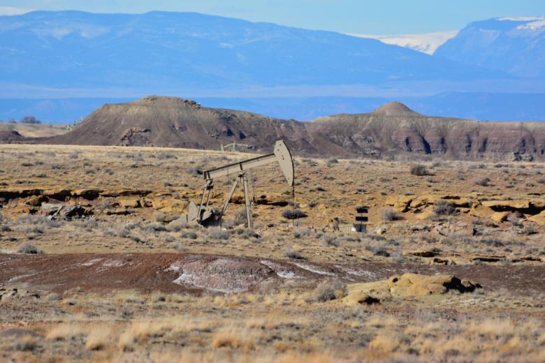 A solitary pump jack or oil well structure stands in a wide, open desert landscape. The terrain is flat and arid with sparse vegetation, scattered rock formations, and low hills or mesas in the distance under a pale sky.