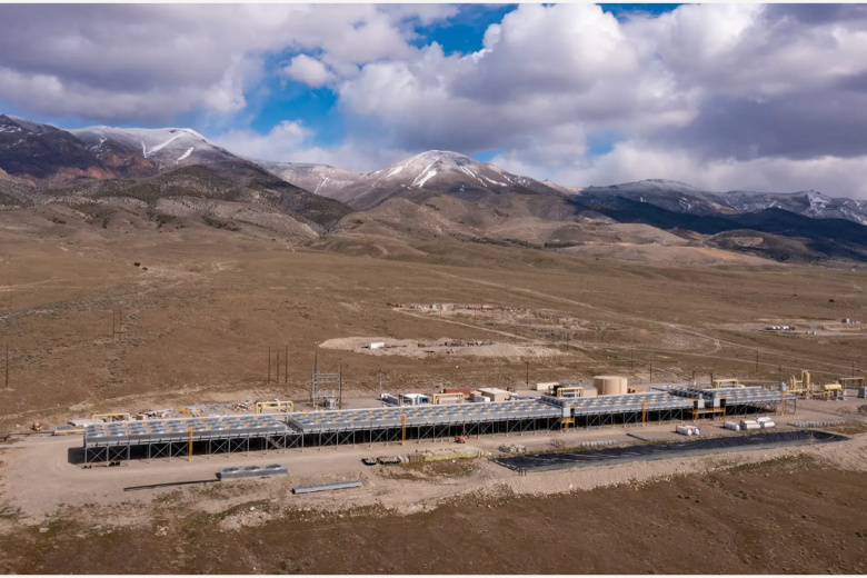 A landscape scene with brown hills and a partly cloudy sky. The foreground is taken up by a long black low-slung structure, a geothermal power plant.