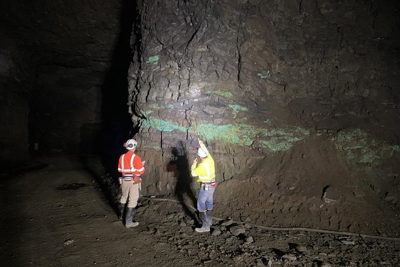 Two miners wearing safety gear inspect a rocky underground wall with green mineral deposits illuminated by a flashlight.