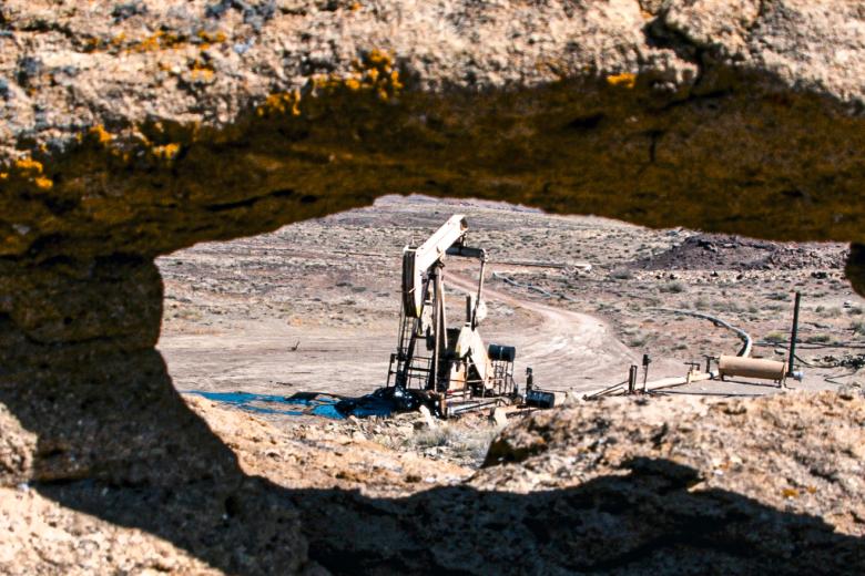 A desert oil pumpjack stands in an open, arid landscape, viewed through a natural opening in the surrounding rock formation. The pumpjack and equipment are framed by rugged stone in the foreground, emphasizing the contrast between industrial activity and the barren environment.