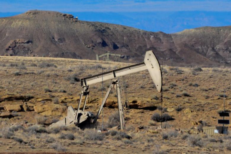 An oil pumpjack operates in a dry desert landscape with sparse vegetation, rocky outcrops, and distant mountains in the background.