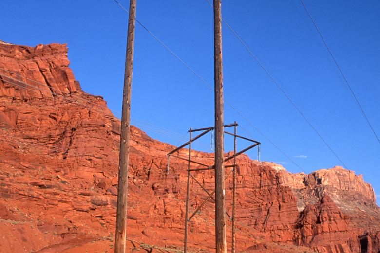 Wooden utility poles stand in a stark desert landscape against a deep blue sky. Behind the towers, massive, layered red rock formations and cliffs rise from the sandy, sun-drenched canyon floor.