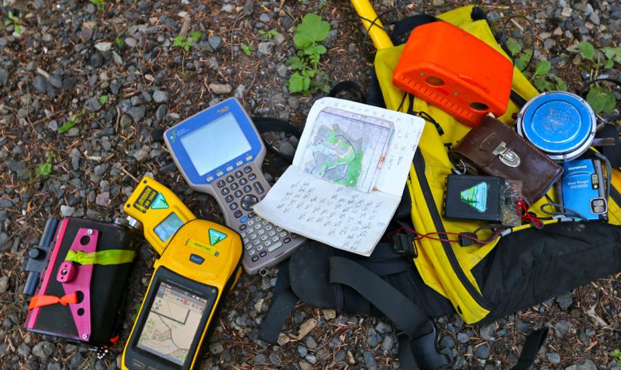 A collection of field equipment is spread out on rocky ground, including handheld GPS units, a rugged data logger, notebooks with maps and handwritten notes, a compass, a camera, and other survey tools placed beside a yellow backpack.