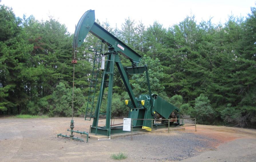 A green oil pump jack stands in a gravel clearing, surrounded by dense trees under a partly cloudy sky.