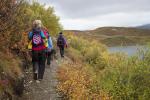 A group of hikers walks single file along a narrow dirt trail through an autumn landscape. They wear backpacks and colorful jackets in red, blue, and purple; the lead hiker carries a child in a pink backpack carrier. The trail is lined with shrubs in fall colors of yellow, orange, and red, with a lake and rolling hills visible under an overcast sky in the background.