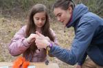 A young girl and an adult outdoors, focusing on a hands-on activity involving small objects on a wooden table.