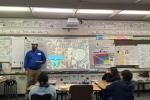 A photo of a man standing in front of students in an elementary school classroom. A display behind him says "Archaeology."
