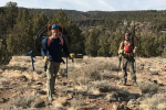 Two Caucasian men in hiking clothes with large rolls of wire and stakes strapped to their backs smiling for the camera outdoors. 