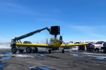 A yellow airplane on tarmac with a snowy backdrop, a large crane holding a box overhead