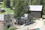 A photo showing several historic wooden buildings with a dirt road going through the town. Wood ramps lead to the entrance of each building. Two large pine trees flank the central building. 