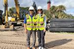 Two people in safety vests stand on a sandy site with heavy machinery and a lighthouse in the background.