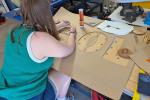 A young woman cuts letters out of a large brown piece of cardboard to make a giant stencil