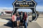 Four people stand in front of a large Route 66 Kingman roadside sign with a vintage vehicle display behind them on a sunny day.
