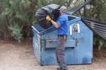 A man throws a bag of trash into a blue dumpster.