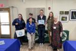 Group of six people standing indoors in front of tables with Federal Junior Duck Stamp banners, one person holding a duck painting.