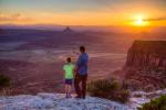 A father and daughter stand on an overlook with landscape features in the distance. 