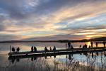People sitting and standing on a dock at sunrise overlooking calm lake waters with colorful clouds reflecting on the surface.