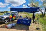 Outdoor event booth under a blue canopy with a colorful prize wheel and informational table near a silver SUV in a grassy field.