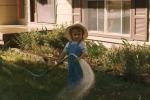 A small child standing in a yard, wearing overalls and a wide-brimmed hat, holding a garden hose that sprays water across the grass near a house.