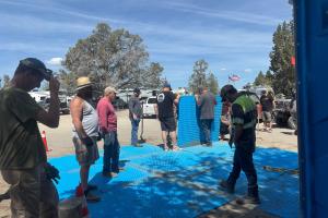 Group of people working outdoors on installing large blue interlocking panels on sandy ground near parked vehicles and trailers, with trees and flags visible in the background under a clear sky.